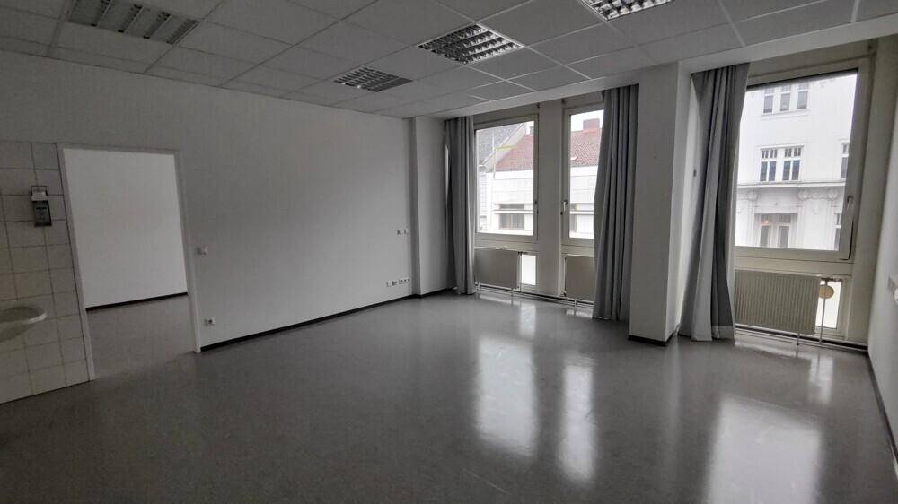 Heller Büroraum mit großen Fenstern, Heizkörpern und einem Waschbeckenbereich an der Seite.
