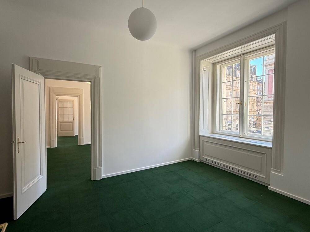 Helles Büro mit grünem Teppichboden, hohen Decken und einem großen Fenster mit Blick nach draußen.
