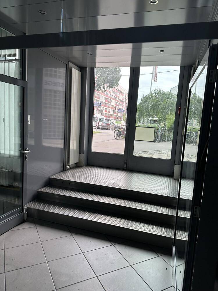 Eingangsbereich mit gefliestem Boden, Metallstufen und Glastüren, die zur Straße führen, mit Blick auf die Umgebung.