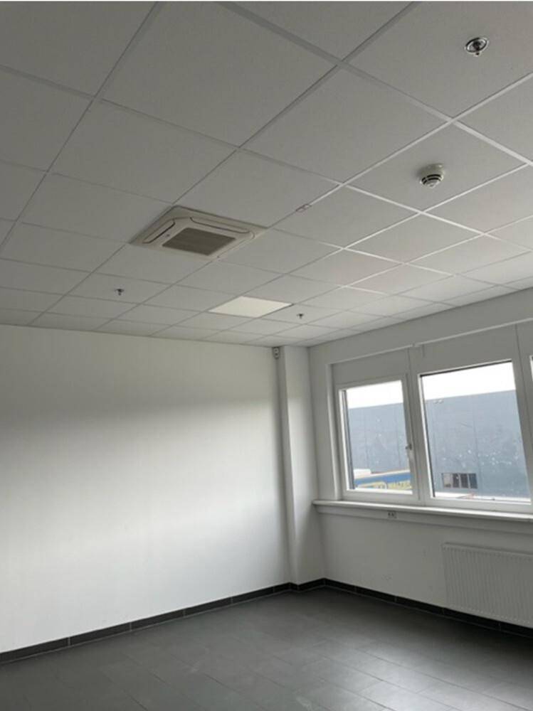 Leerer Büroraum mit weißen Wänden, grauen Fliesen und einem Fenster mit Blick auf ein Industriegebäude.