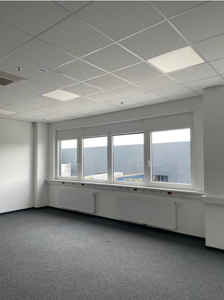 Heller Büroraum mit grauem Teppichboden, großen Fenstern und Heizkörpern unter den Fensterbänken.