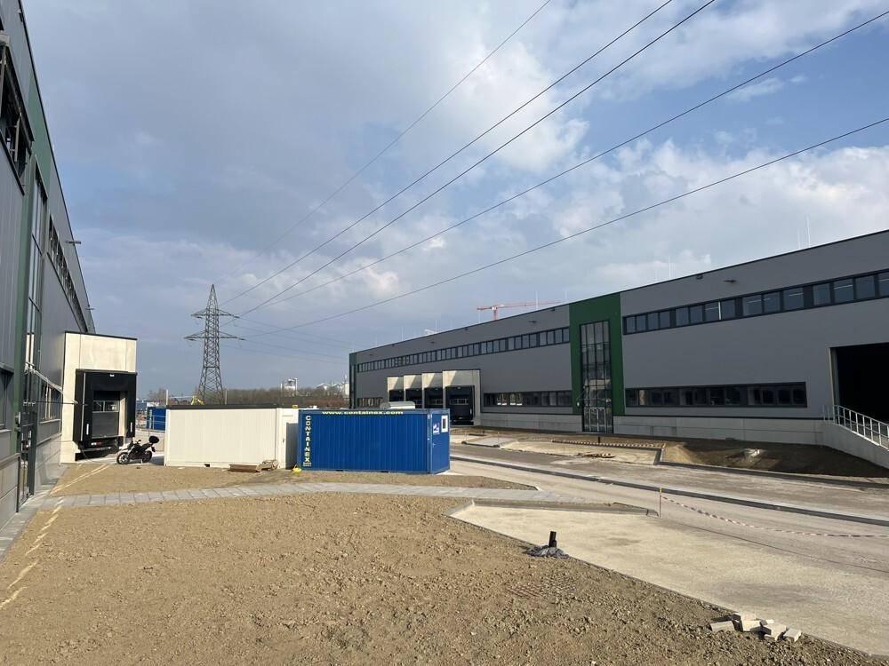 Die Außenseite des Industriegebäudes mit Laderampen, grüner Fassade und Baucontainern unter Stromleitungen.