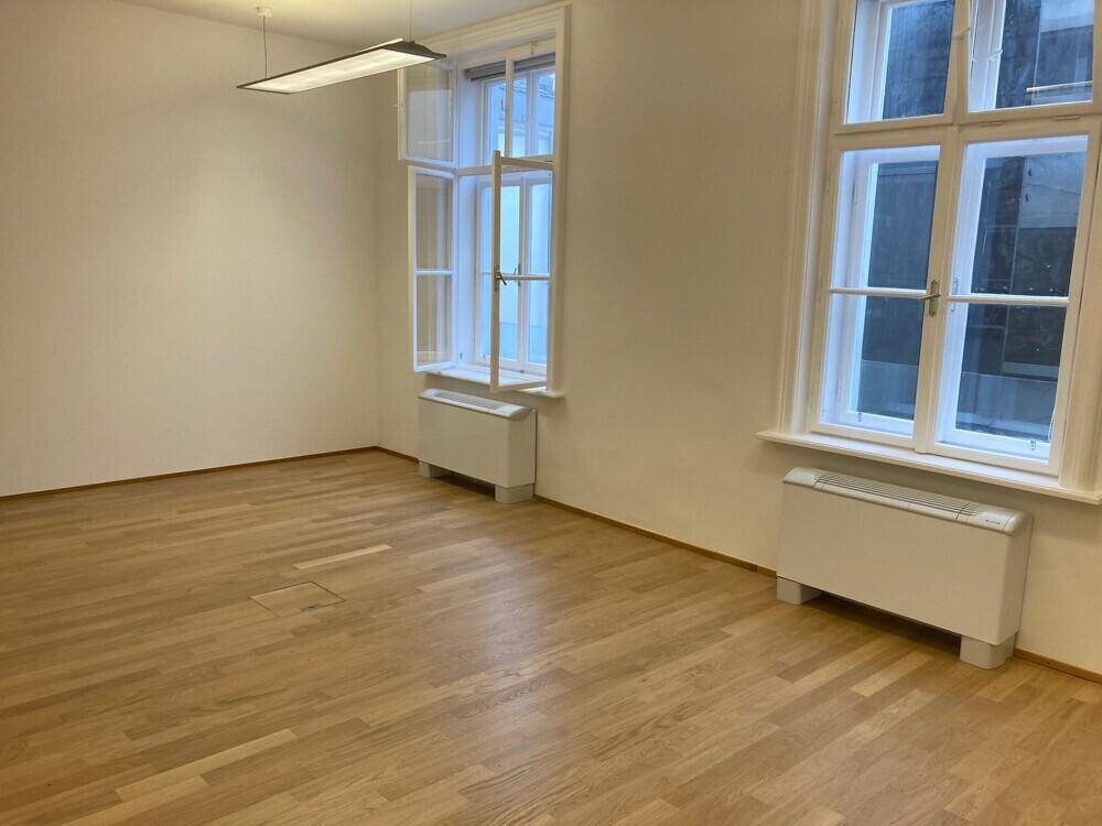Geräumiges Büro mit Holzboden und zwei geöffneten Fenstern, die frische Luft hereinlassen.