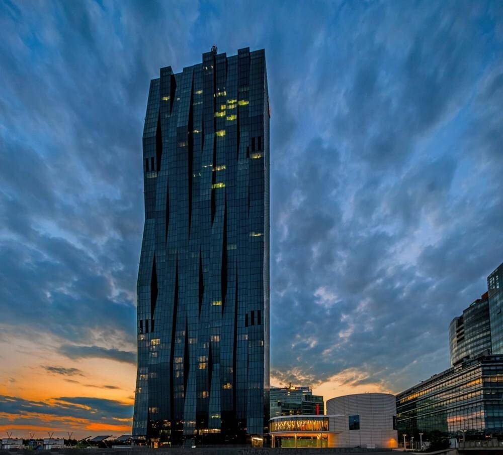 Dramatische Ansicht des modernen Wolkenkratzers bei Sonnenuntergang mit beleuchteten Fenstern und dunklem Himmel.