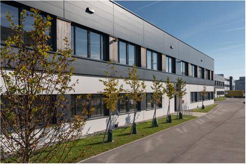 Seitenansicht eines modernen Industriegebäudes mit großen Fenstern und jungen Bäumen entlang der Fassade.