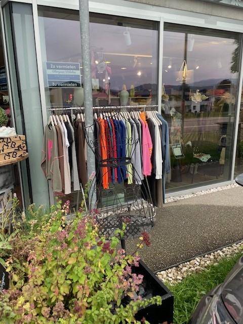 Außenansicht eines Geschäfts mit Kleiderständer vor dem Schaufenster und "Zu vermieten"-Schildern im Inneren.