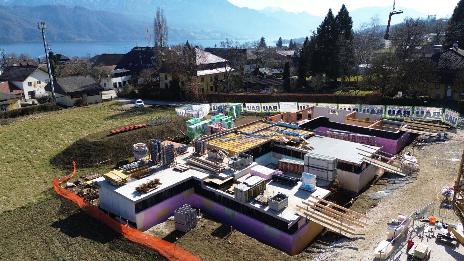 Luftaufnahme einer Baustelle mit Fundamenten und beginnendem Rohbau in Seenähe.