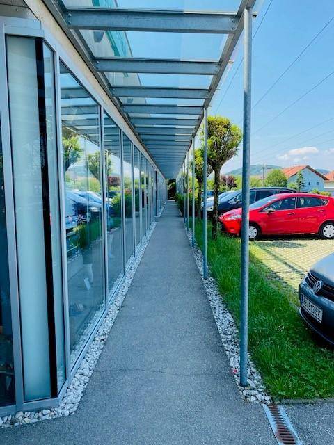 Überdachter Gehweg entlang einer Reihe von Ladenfronten mit großen Glasfenstern und Parkplätzen im Hintergrund.