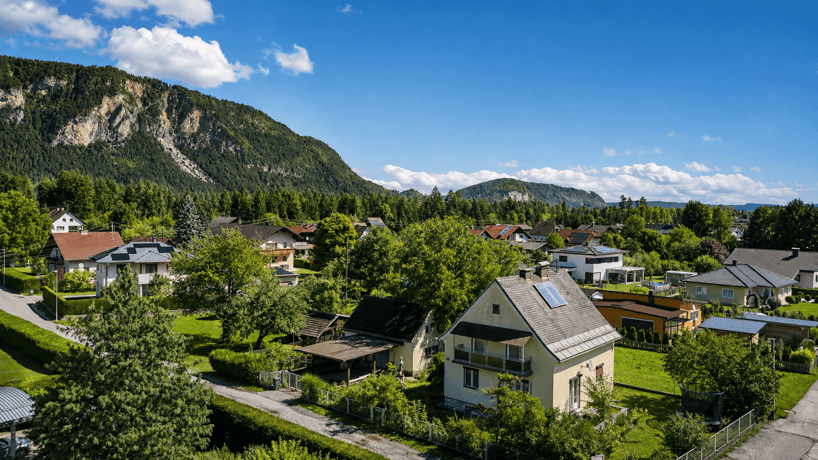 Luftaufnahme einer Wohnsiedlung mit Einfamilienhäusern, umgeben von grünen Wäldern und Bergen.