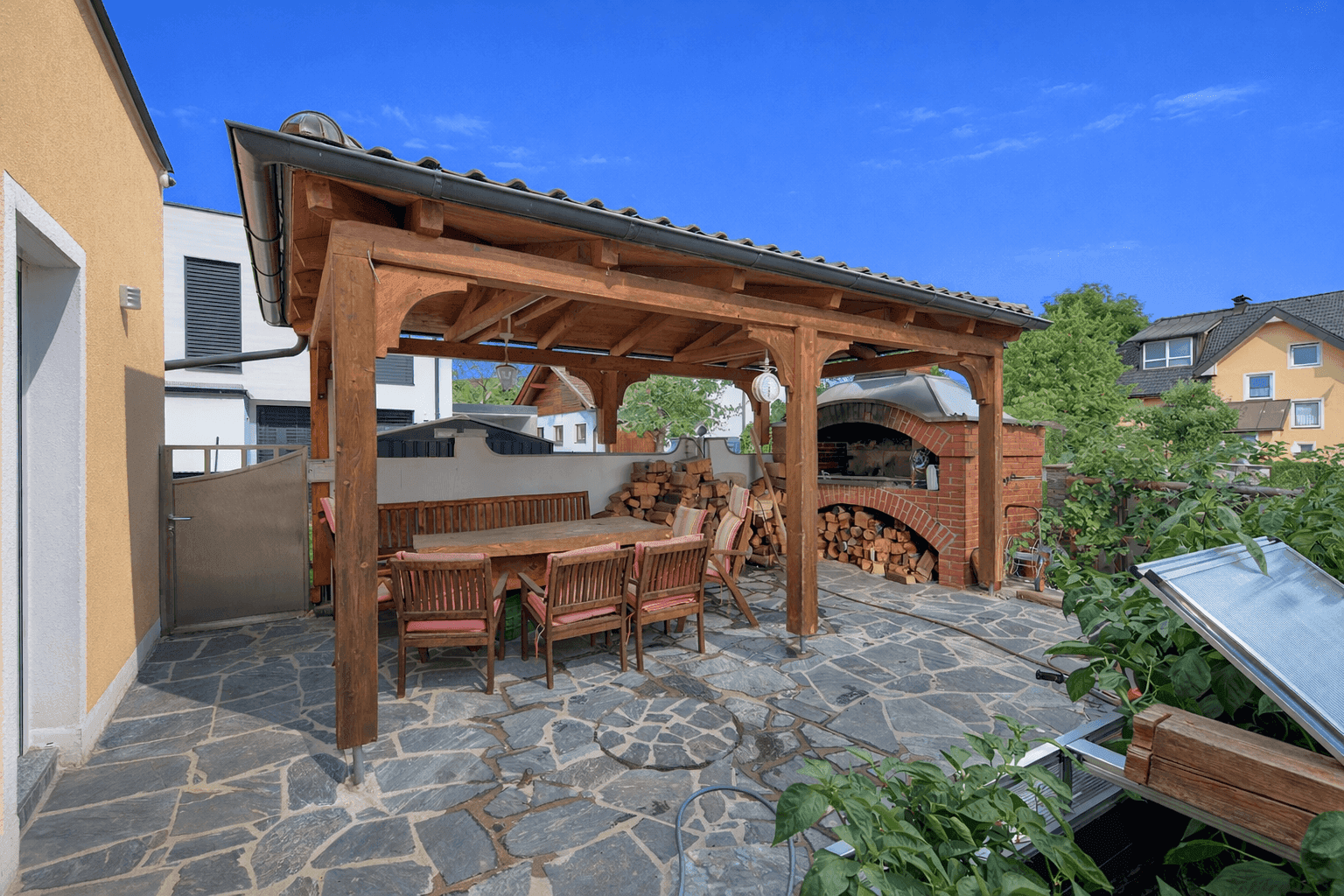 Rustikaler Essbereich im Freien mit Holzpergola und großem gemauerten Pizzaofen.