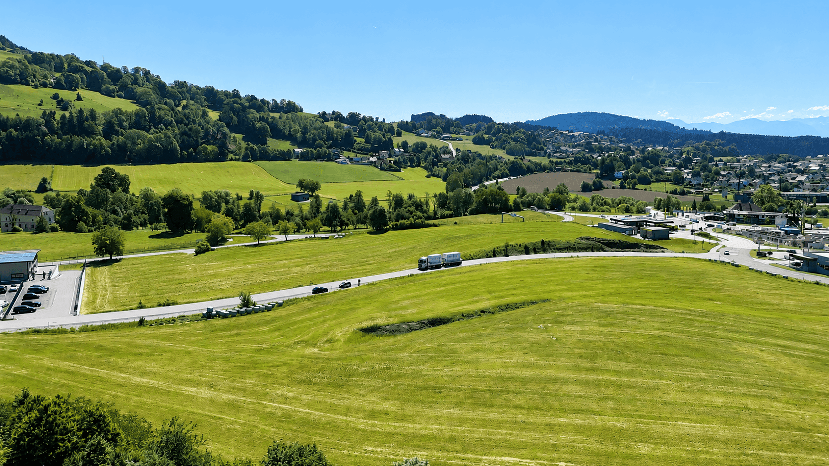 Luftaufnahme von weiten grünen Feldern mit einer Straße, Gebäuden und bewaldeten Hügeln im Hintergrund.