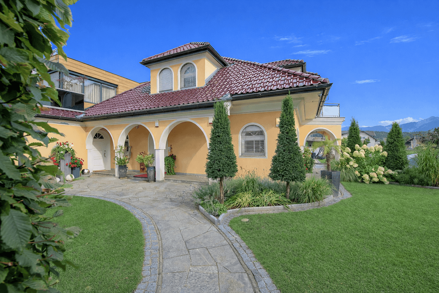 Elegante Villa mit gepflegtem Vorgarten, geschwungenem Steinweg und Bergblick im Hintergrund.
