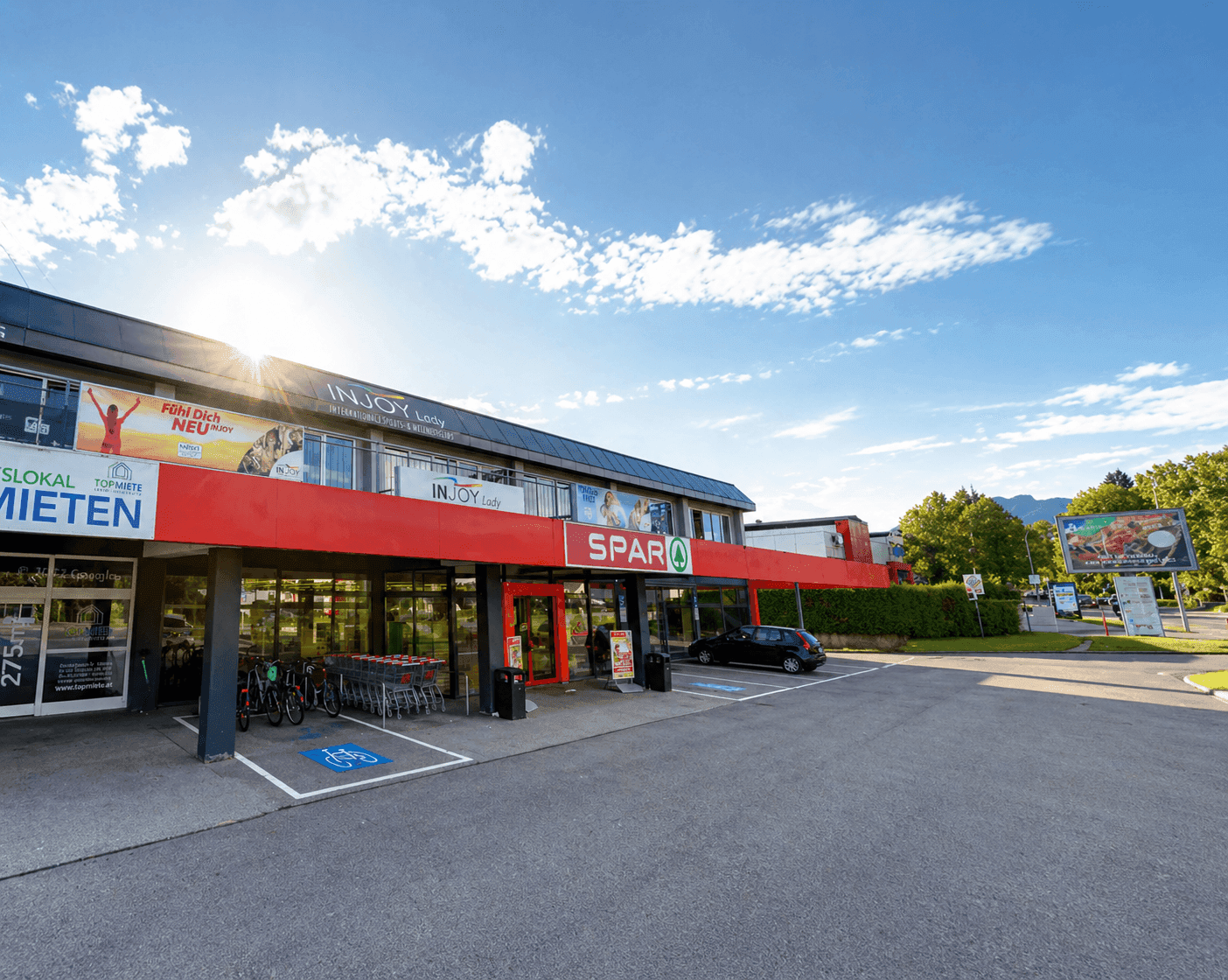 Frontansicht eines Geschäftsgebäudes mit Supermarkt-Eingang und Parkplätzen bei Sonnenschein.