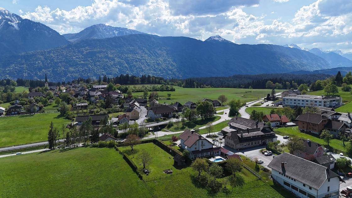 Weitläufige Luftaufnahme eines Dorfes eingebettet in eine grüne Landschaft mit Bergen und einem See in der Ferne.