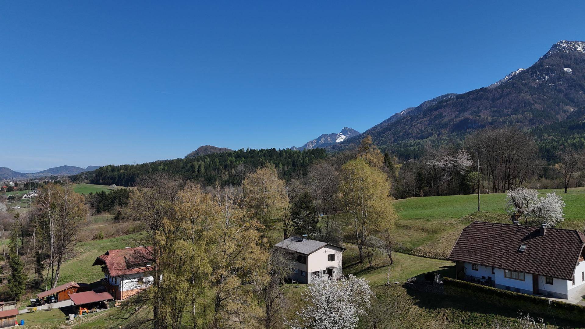 Panoramablick auf mehrere Häuser in einer ländlichen Umgebung mit weiten Feldern und majestätischen Bergen im Hintergrund.