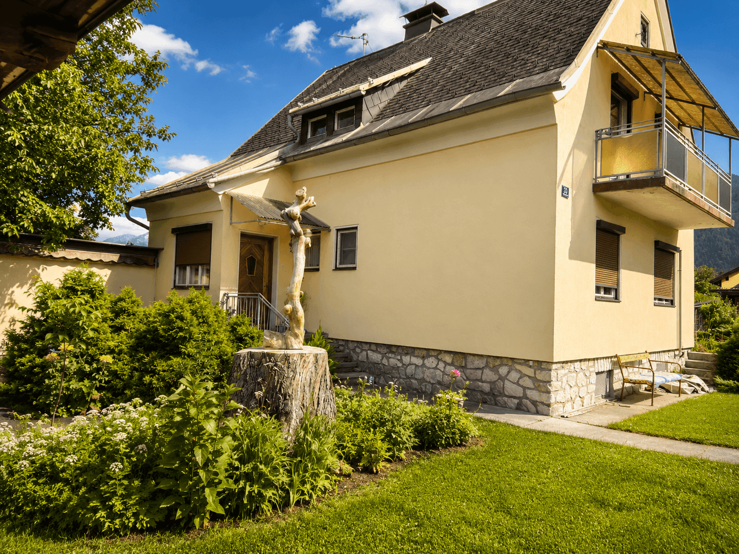 Seitenansicht des gelben Hauses mit gepflegtem Garten, Rasenfläche und einem kleinen Balkon.