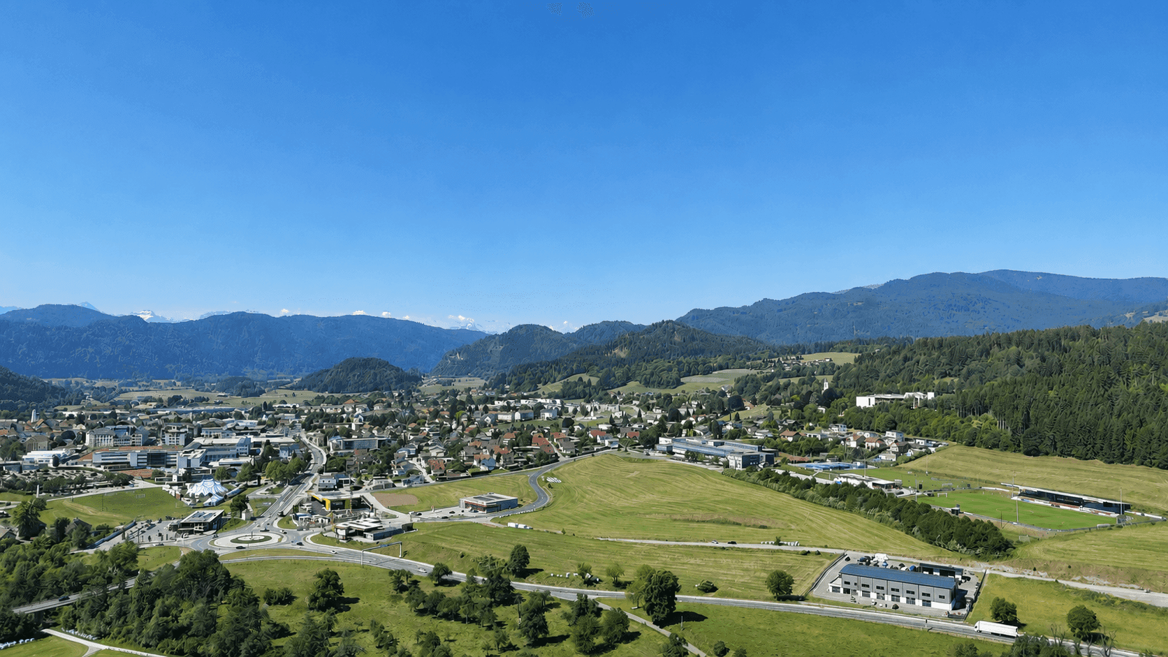 Panoramablick auf eine ländliche Stadt umgeben von grünen Hügeln, Wäldern und fernen Bergen unter blauem Himmel.