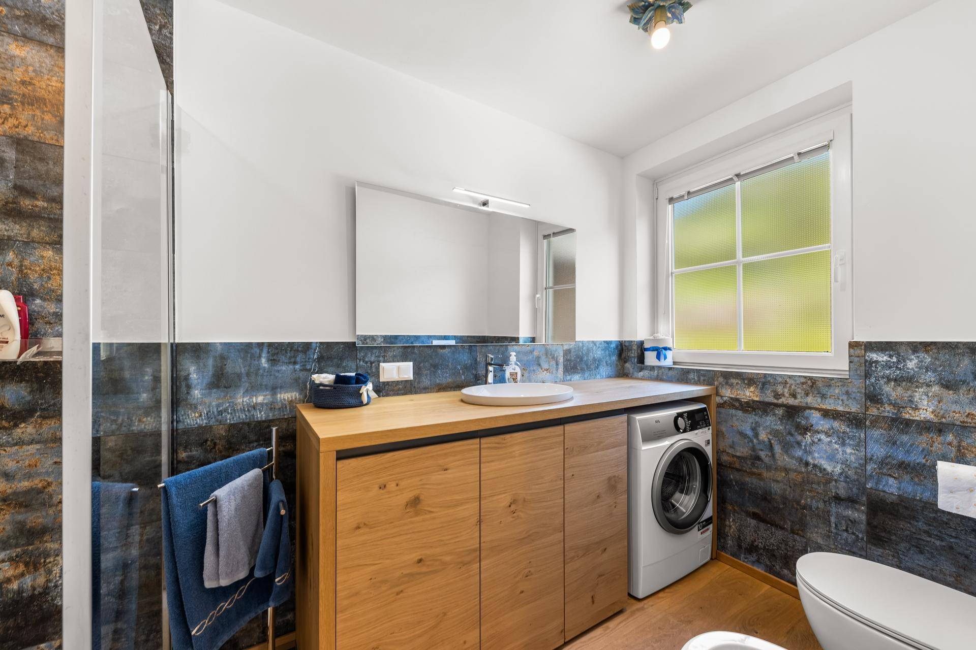 Funktionales Badezimmer mit Waschmaschine, Einzelwaschbecken und Dusche, hell durch ein Fenster.