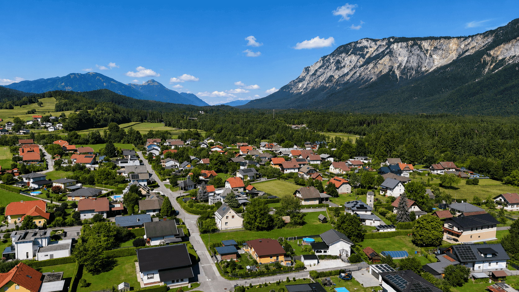 Weitläufige Luftaufnahme einer ländlichen Wohnsiedlung, umgeben von Wäldern und majestätischen Bergen.