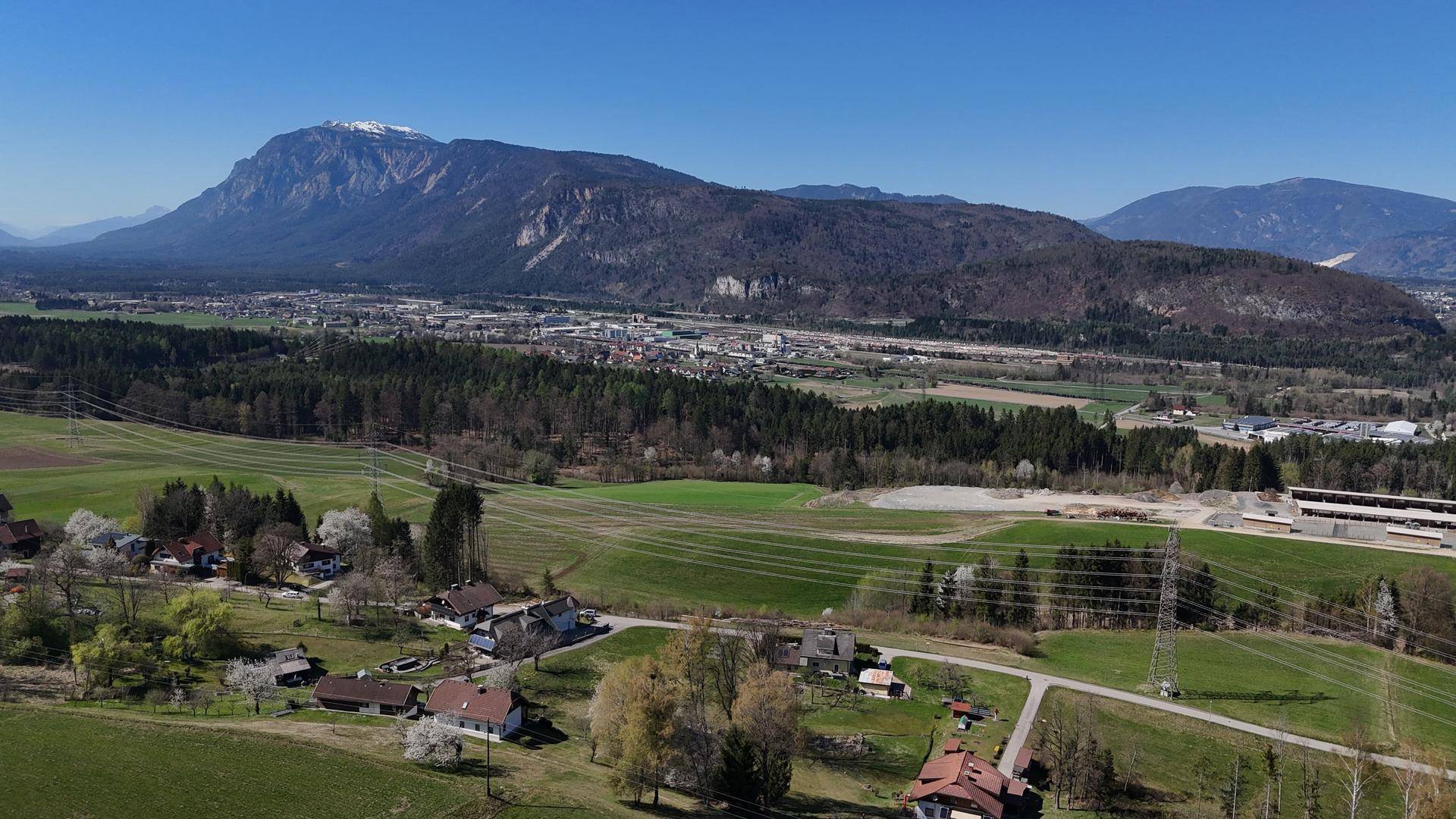 Panoramische Luftaufnahme einer Siedlung mit Feldern und einem großen Berg im Hintergrund unter blauem Himmel.
