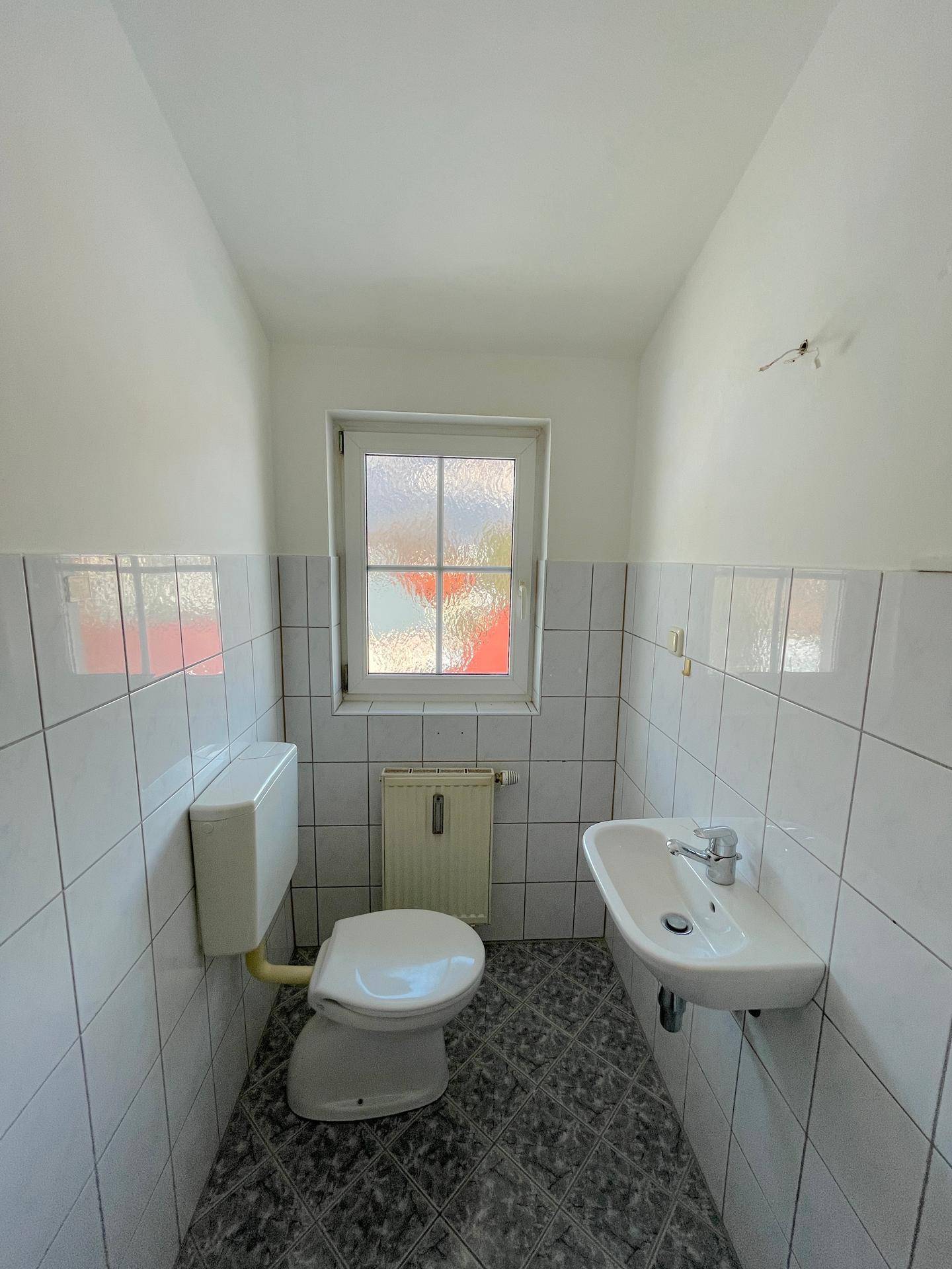 Kleines Gäste-WC mit Fenster, Toilette, Waschbecken und weißen Fliesen an den Wänden.