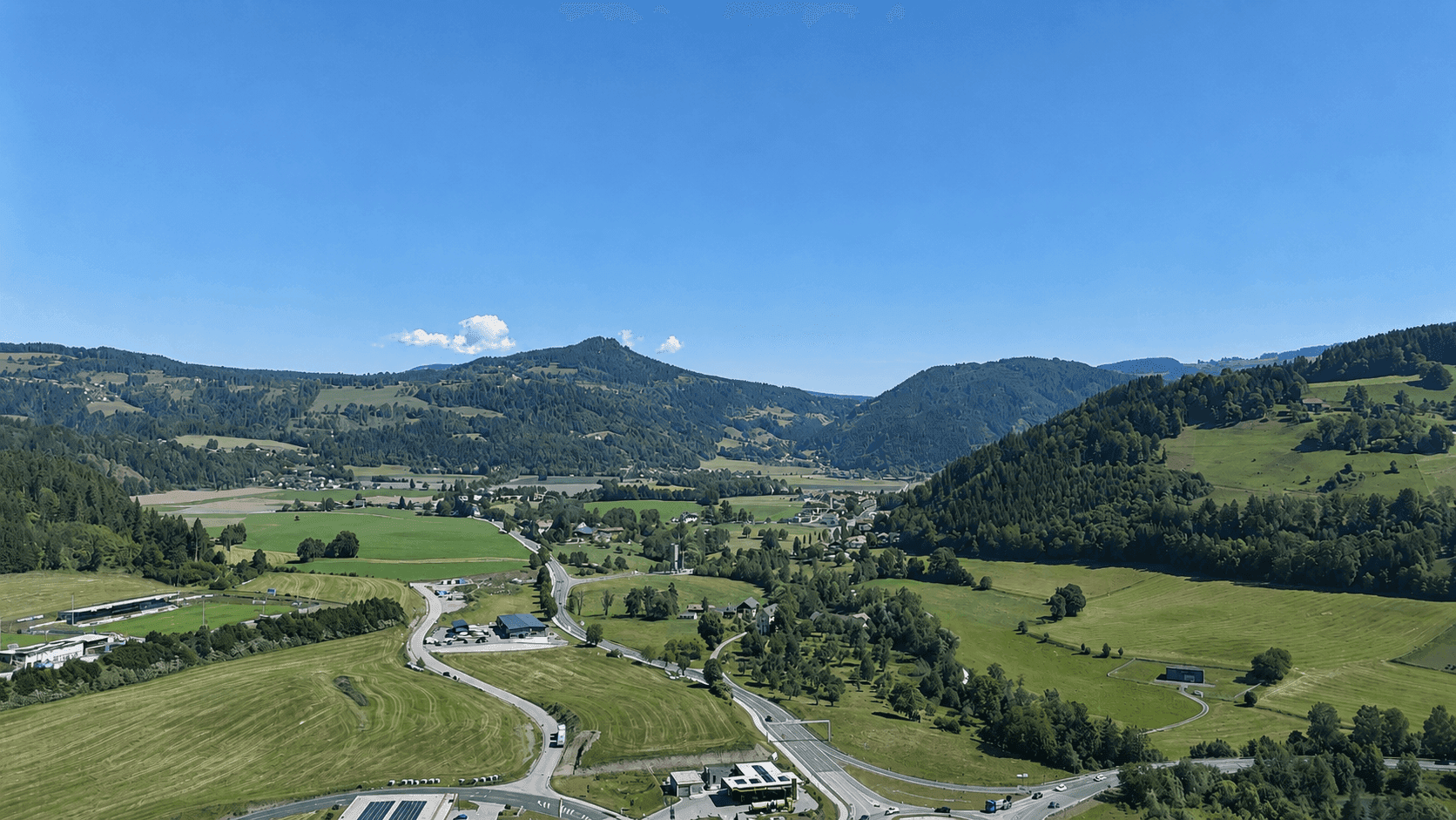 Weitläufiger Blick über ein grünes Tal mit einem Dorf, umgeben von dichten Wäldern und sanften Hügeln.