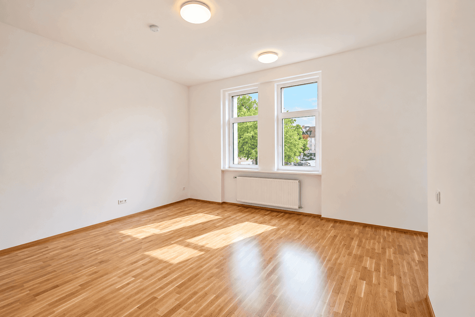Helles Zimmer mit Parkettboden, zwei großen Fenstern und viel Tageslicht.