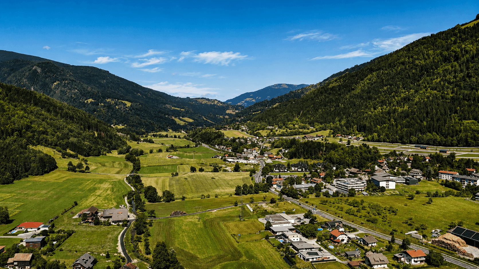 Weitwinkelaufnahme eines Tals mit bewaldeten Bergen, einer Straße und verstreuten Häusern.