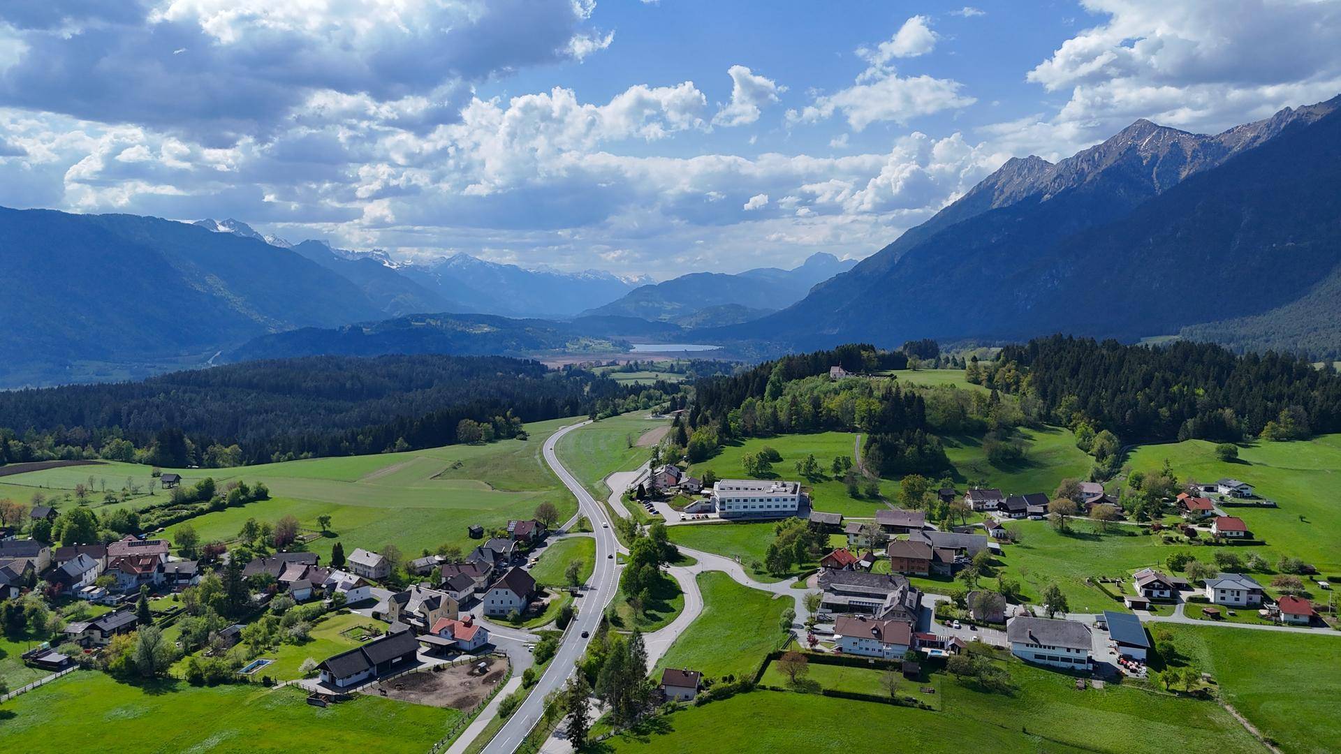 Panoramische Luftaufnahme einer ländlichen Siedlung mit Bergen, Wäldern und einem See in der Ferne.