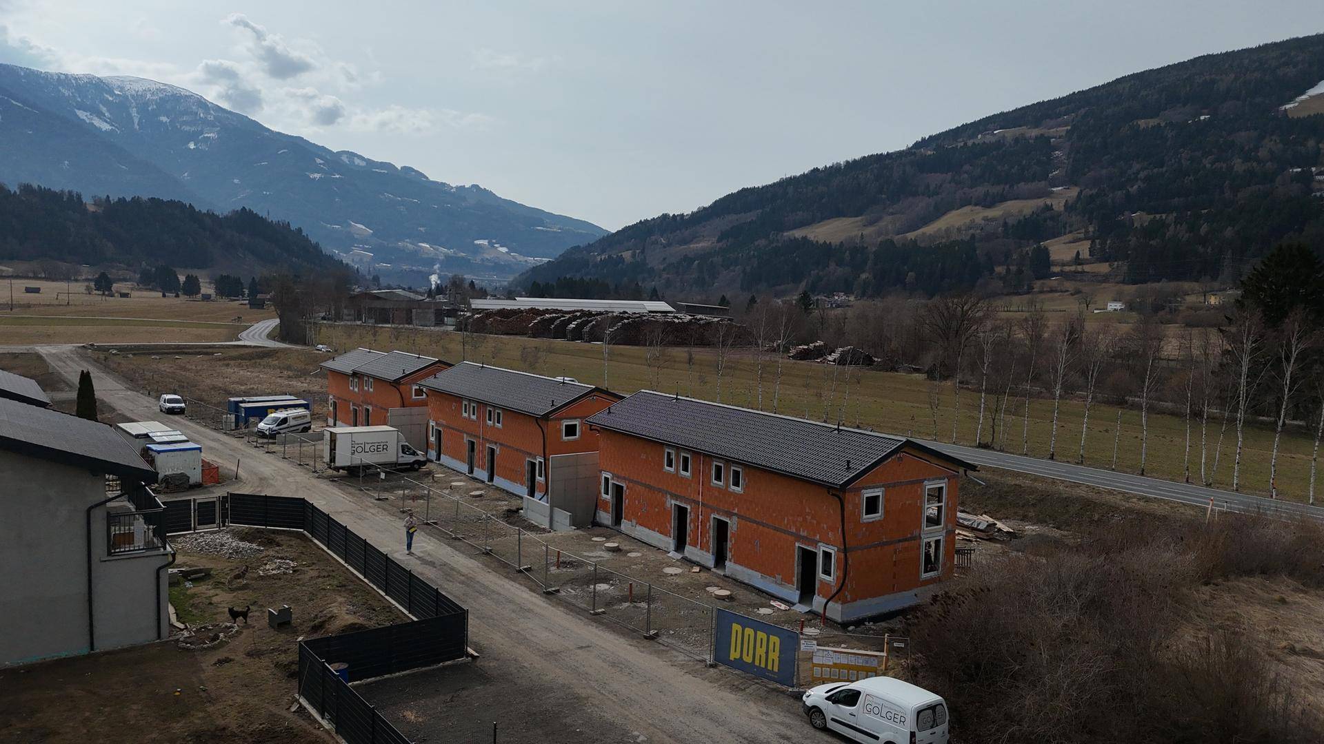 Luftaufnahme der im Bau befindlichen Reihenhäuser entlang einer Straße mit Bergpanorama im Hintergrund.