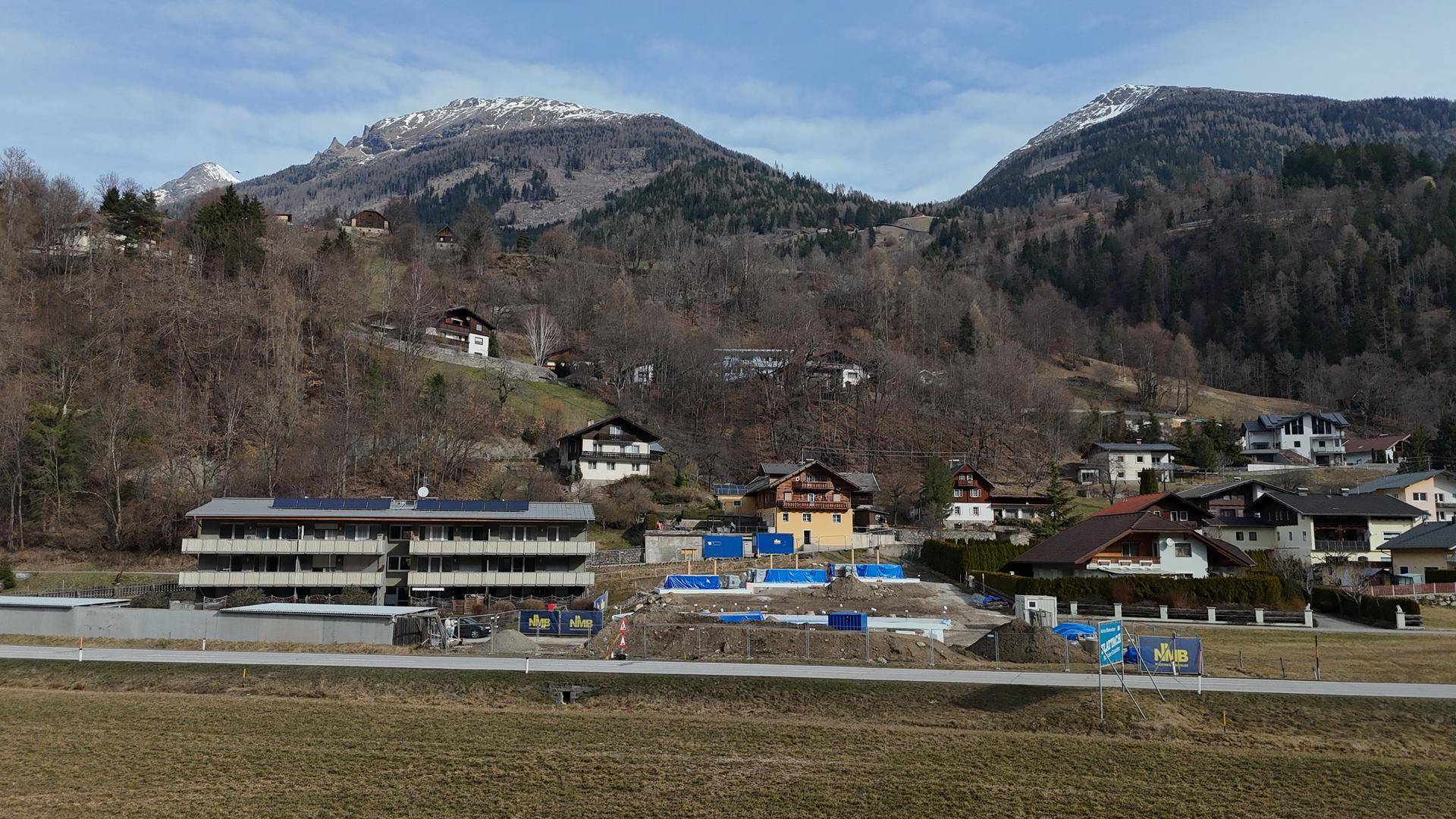 Blick auf eine Baustelle mit bestehenden Gebäuden und einer beeindruckenden Bergkulisse im Hintergrund.