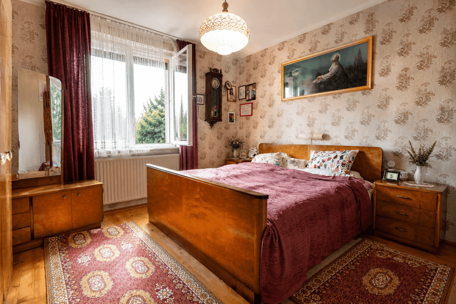 Schlafzimmer mit Doppelbett, Holzmöbeln, gemusterter Tapete und einem Fenster mit Blick ins Grüne.