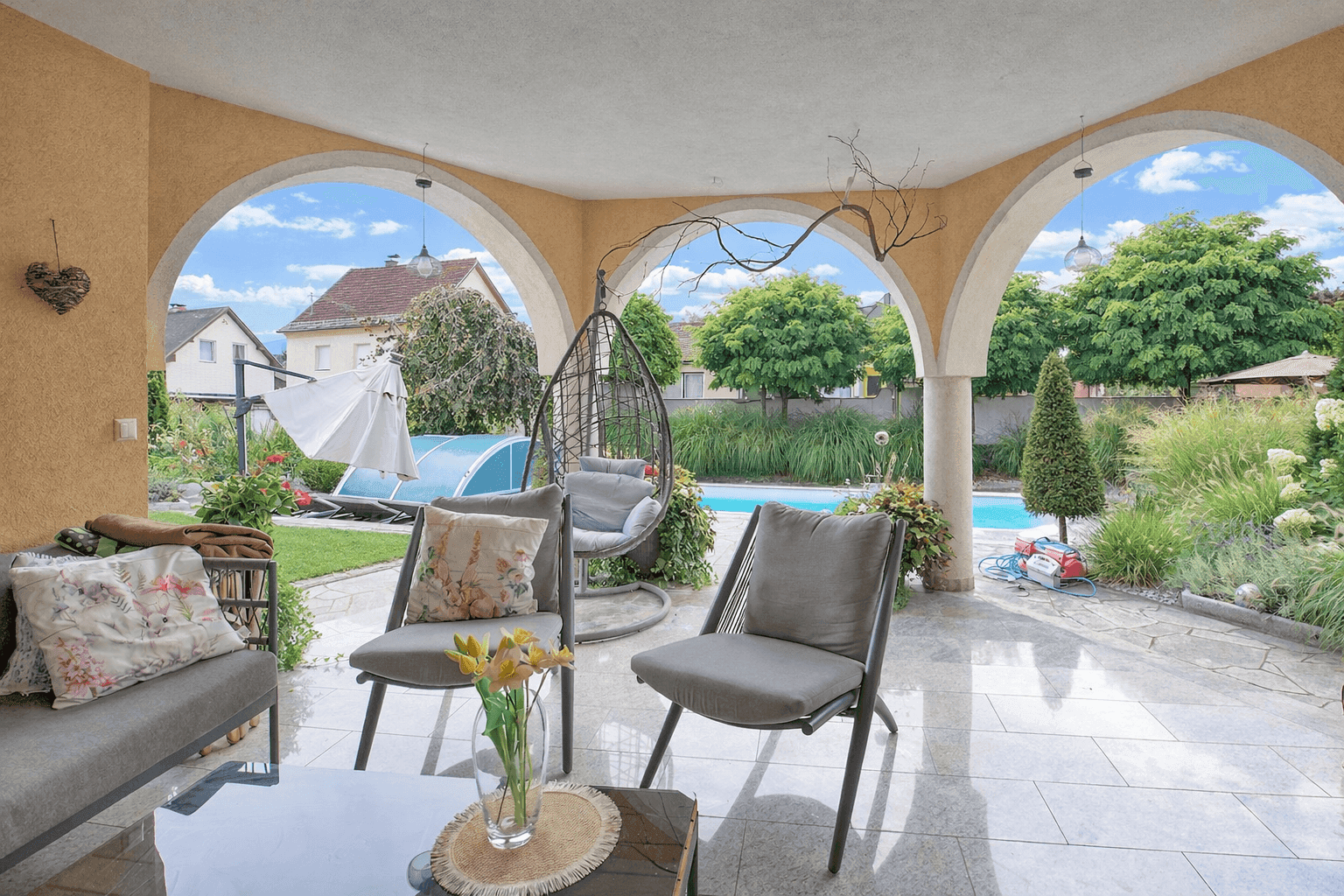 Überdachte Terrasse mit bequemen Sitzgelegenheiten und Blick auf den Pool und Garten.