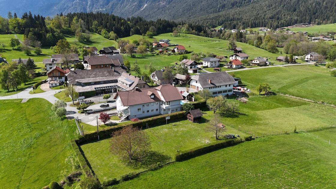 Luftaufnahme eines Hauses in einer grünen, ländlichen Umgebung mit weiteren Gebäuden und Bergen im Hintergrund.