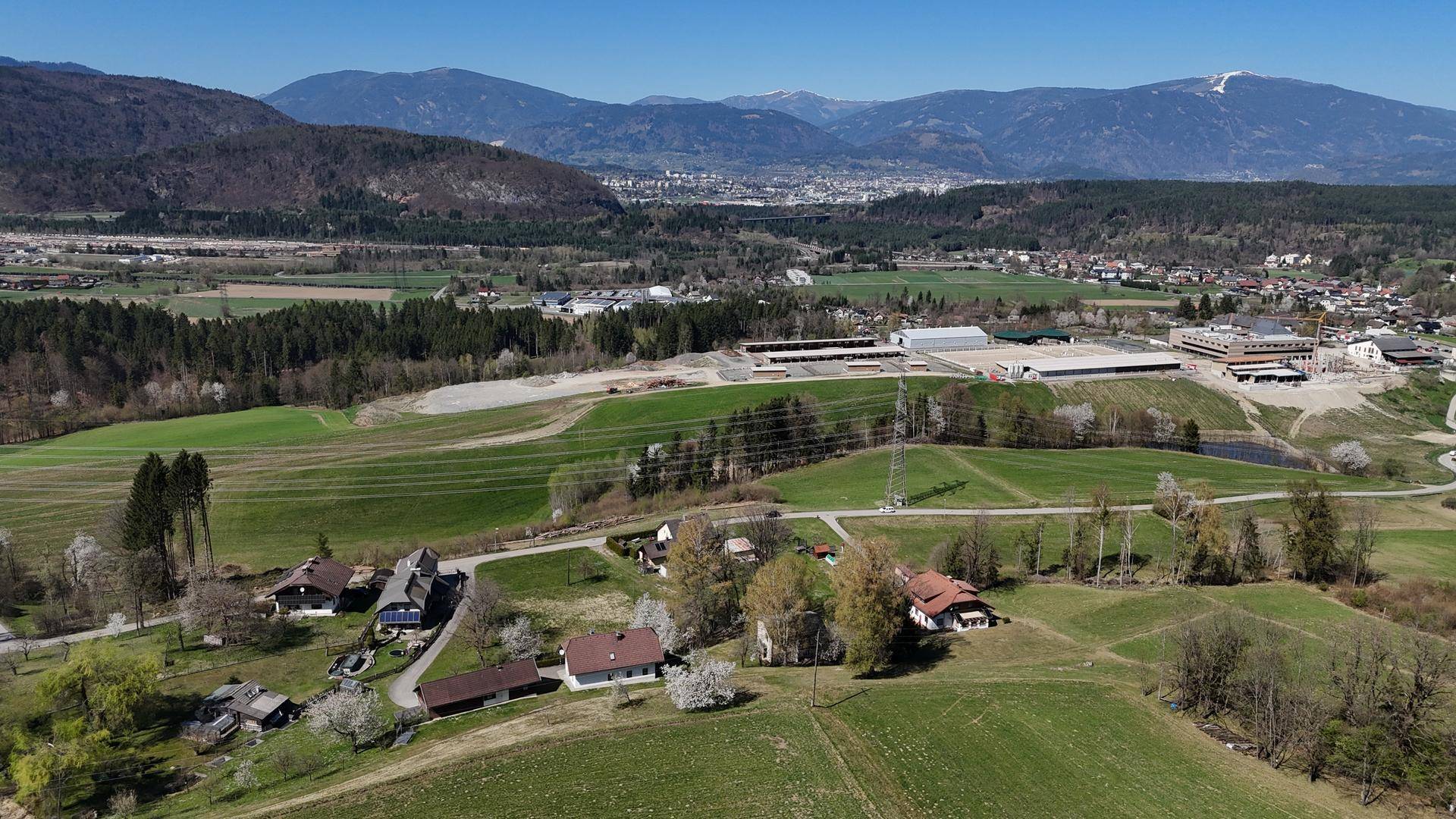 Luftaufnahme einer ländlichen Gegend mit Häusern, Feldern und einem Industriegebiet, umgeben von Bergen.