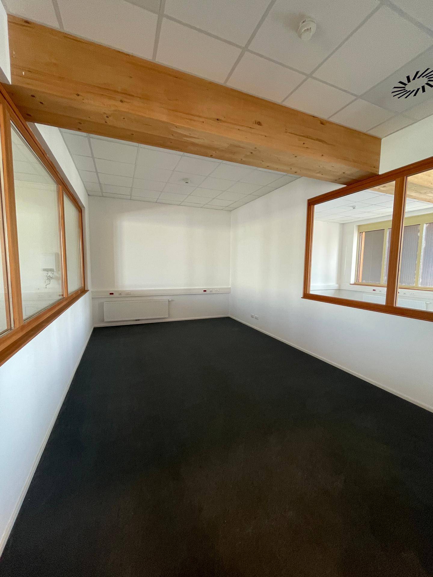 Büro mit dunklem Teppichboden, Holzfenstern und einem Heizkörper, mit Blick in einen Nebenraum.