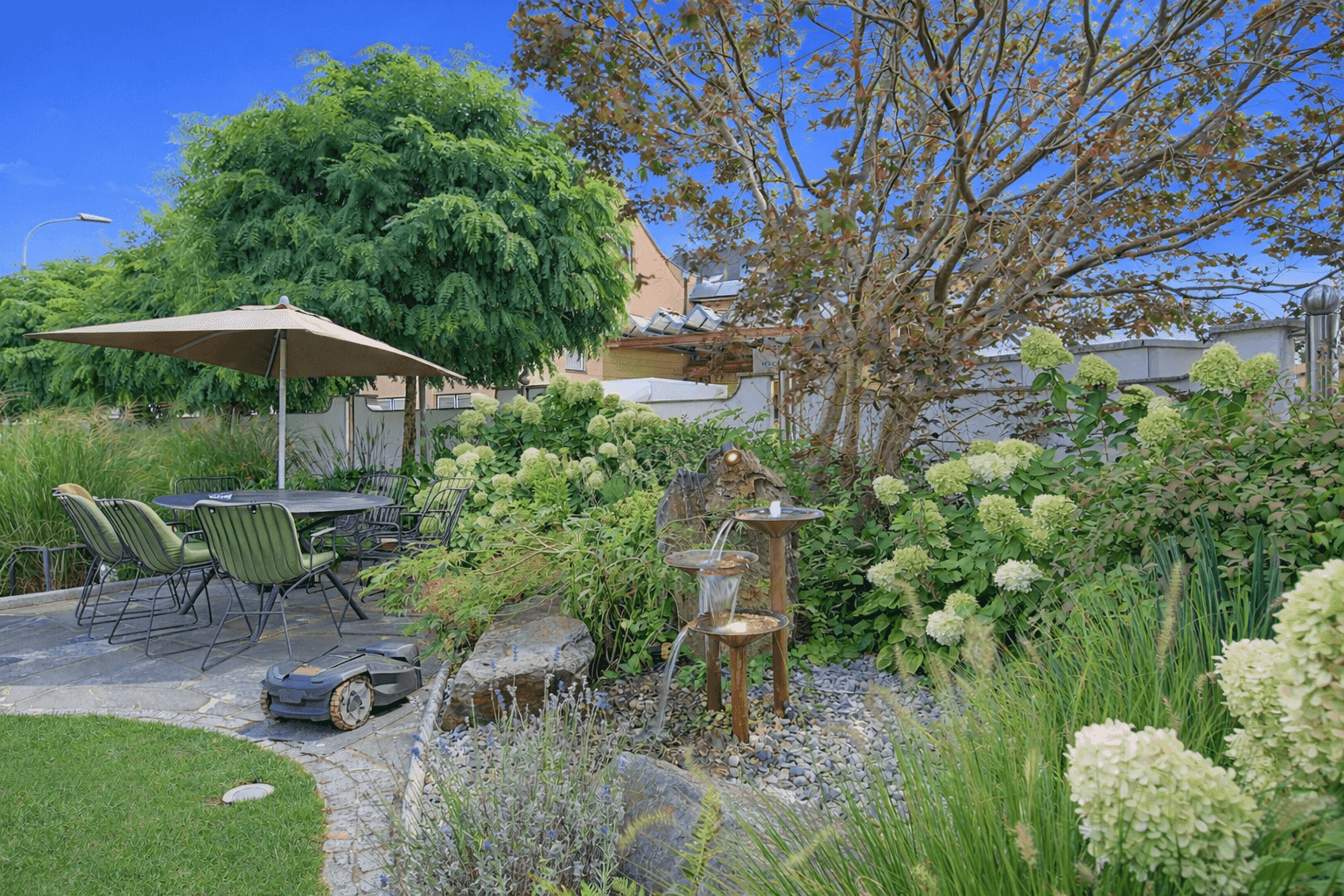 Gepflegter Garten mit Wasserspiel, Rasenroboter und schattigem Essbereich unter einem Sonnenschirm.