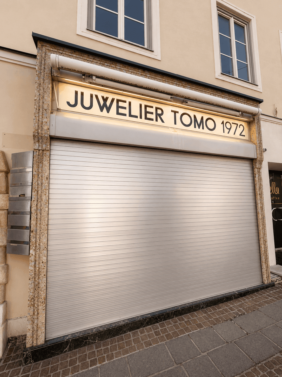Fassade eines Juweliergeschäfts mit geschlossenem Rollladen und beleuchtetem Firmenschild an der Straße.
