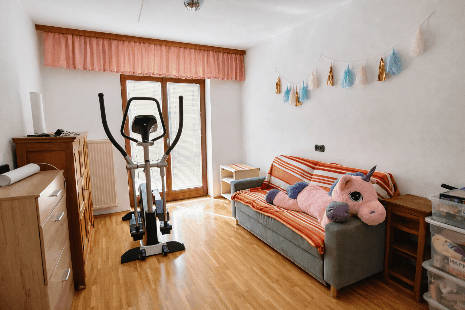 Helles Zimmer mit Holzboden, Sofa und Trainingsgerät, mit Zugang zum Balkon.