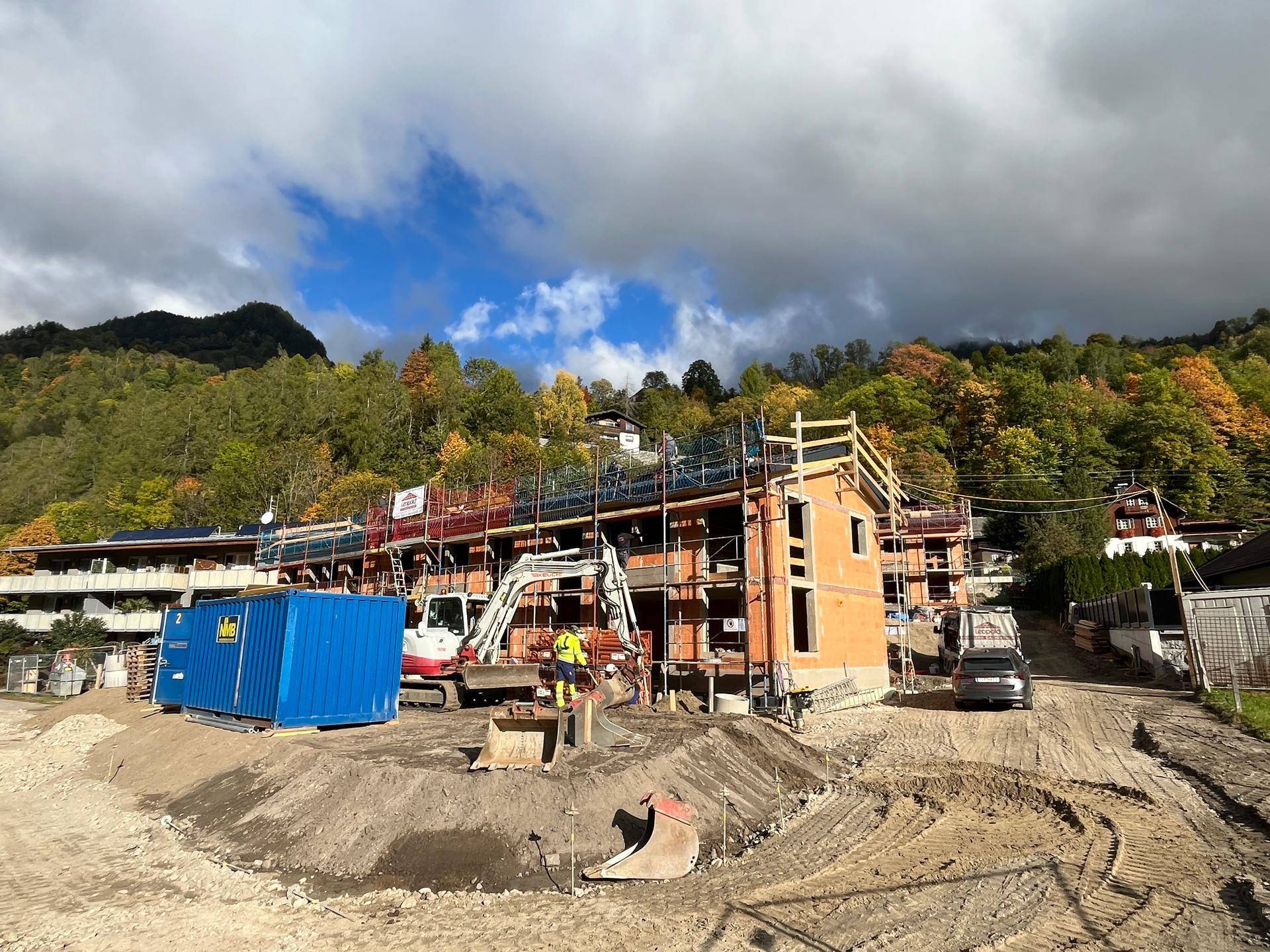 Baustelle eines Neubauprojekts mit Baggern und Containern vor einer malerischen Bergkulisse.