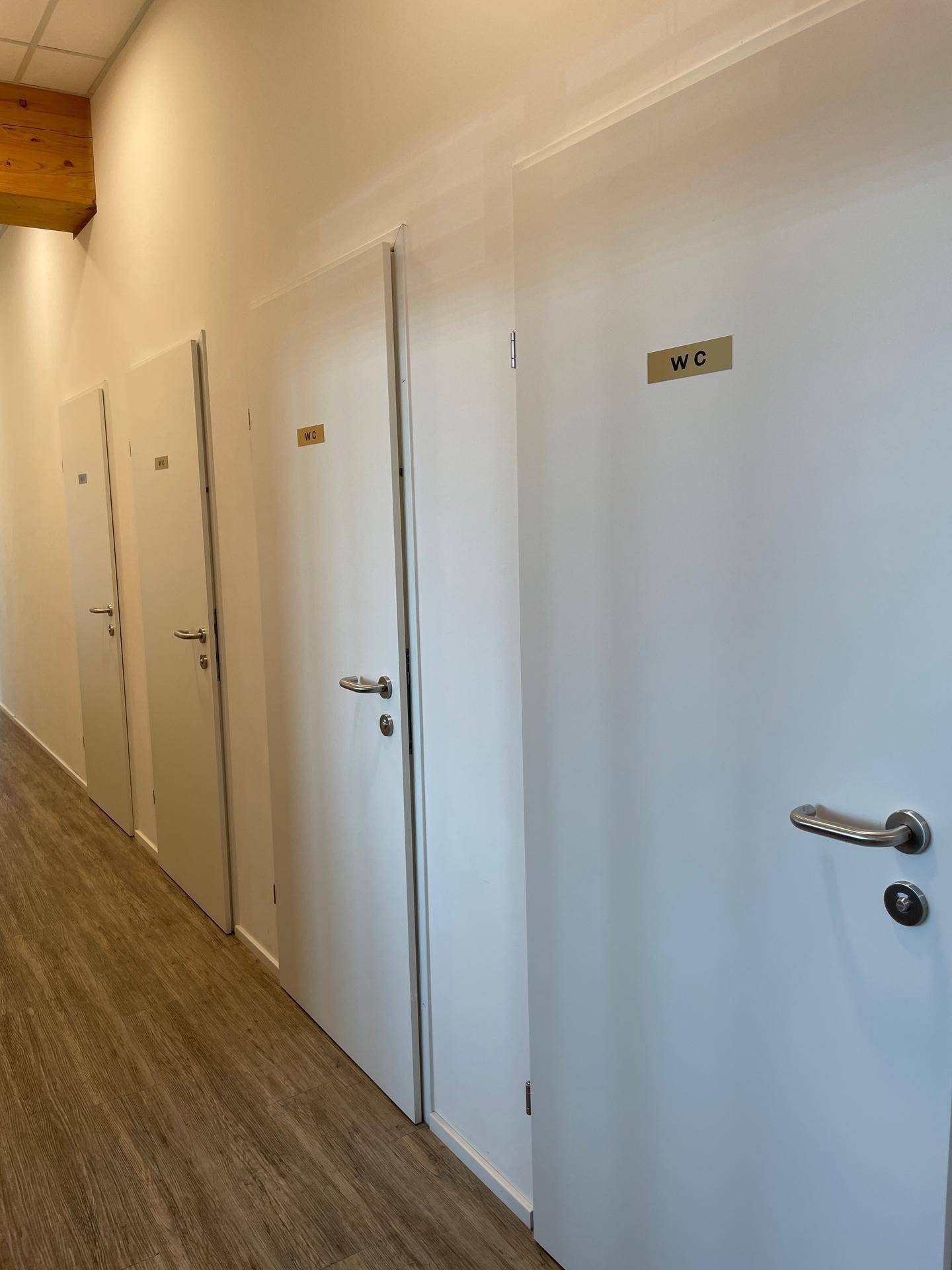 Flur mit mehreren weißen Türen, darunter Toilettenräume, und hellem Bodenbelag.