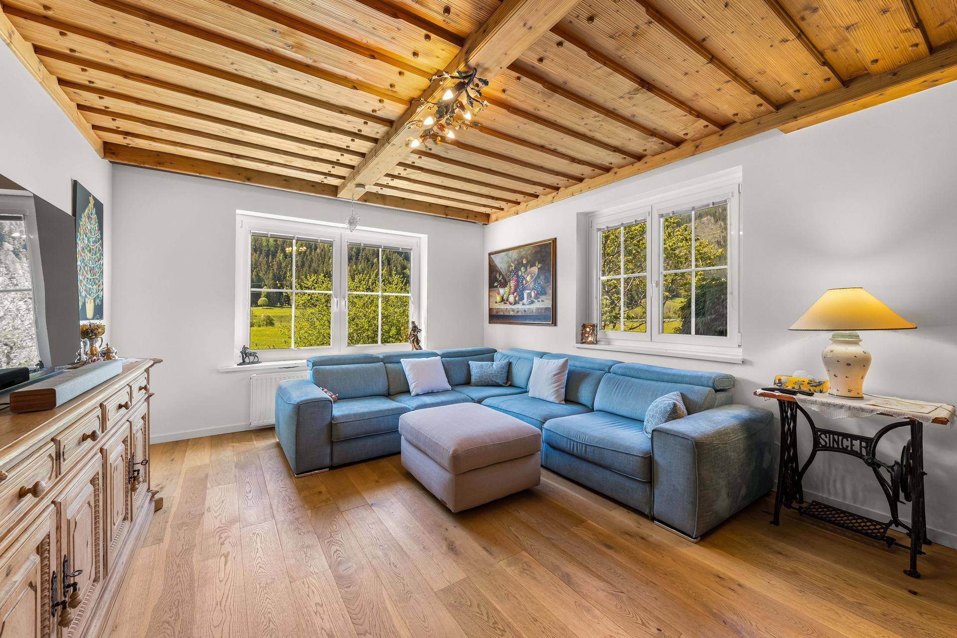 Geräumiges Wohnzimmer mit bequemer blauer Couch, Holzbalkendecke und weitem Blick auf die grüne Landschaft.