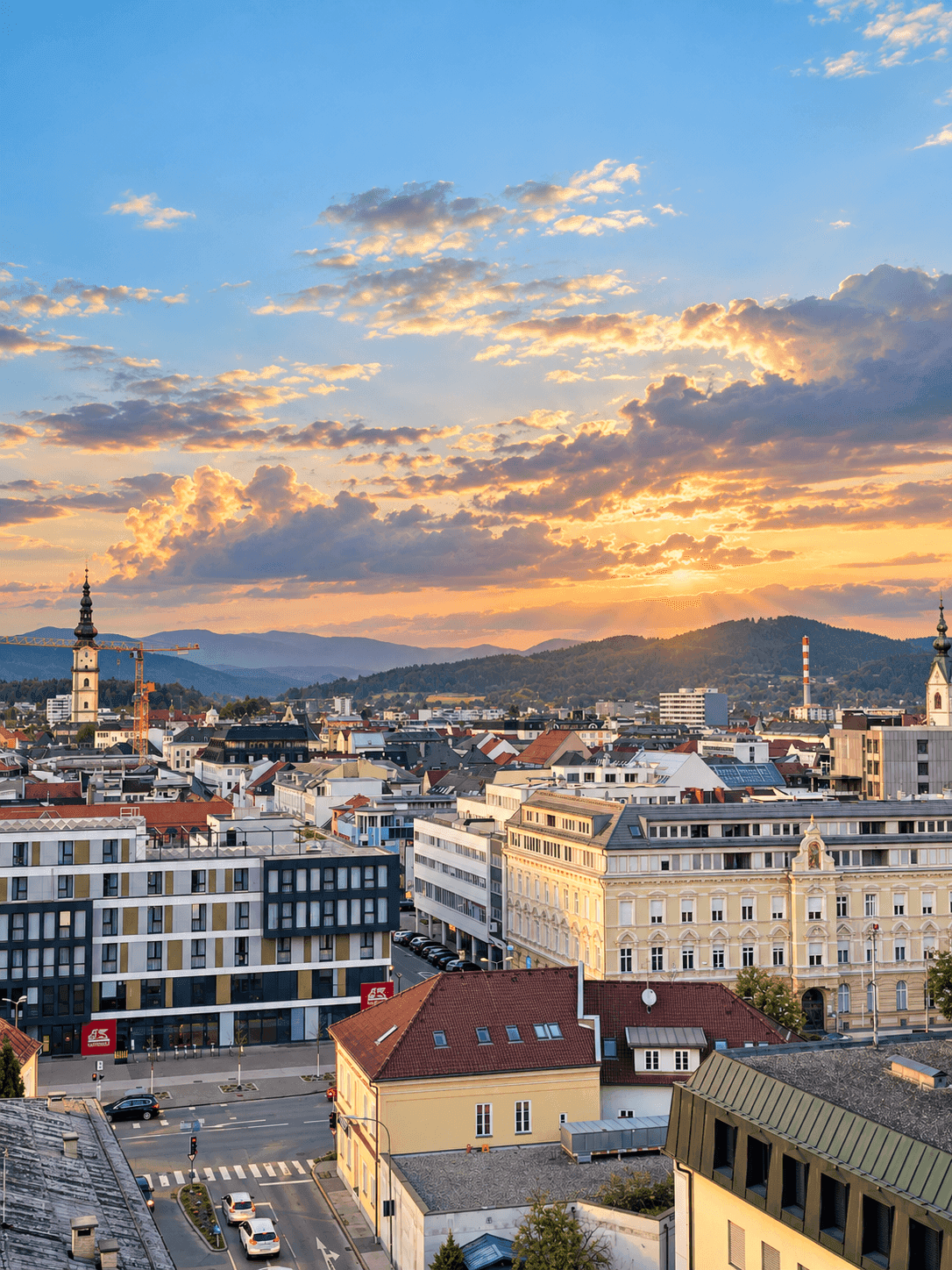 Städtische Skyline bei Sonnenuntergang mit Bergen im Hintergrund und dramatischen Wolken.