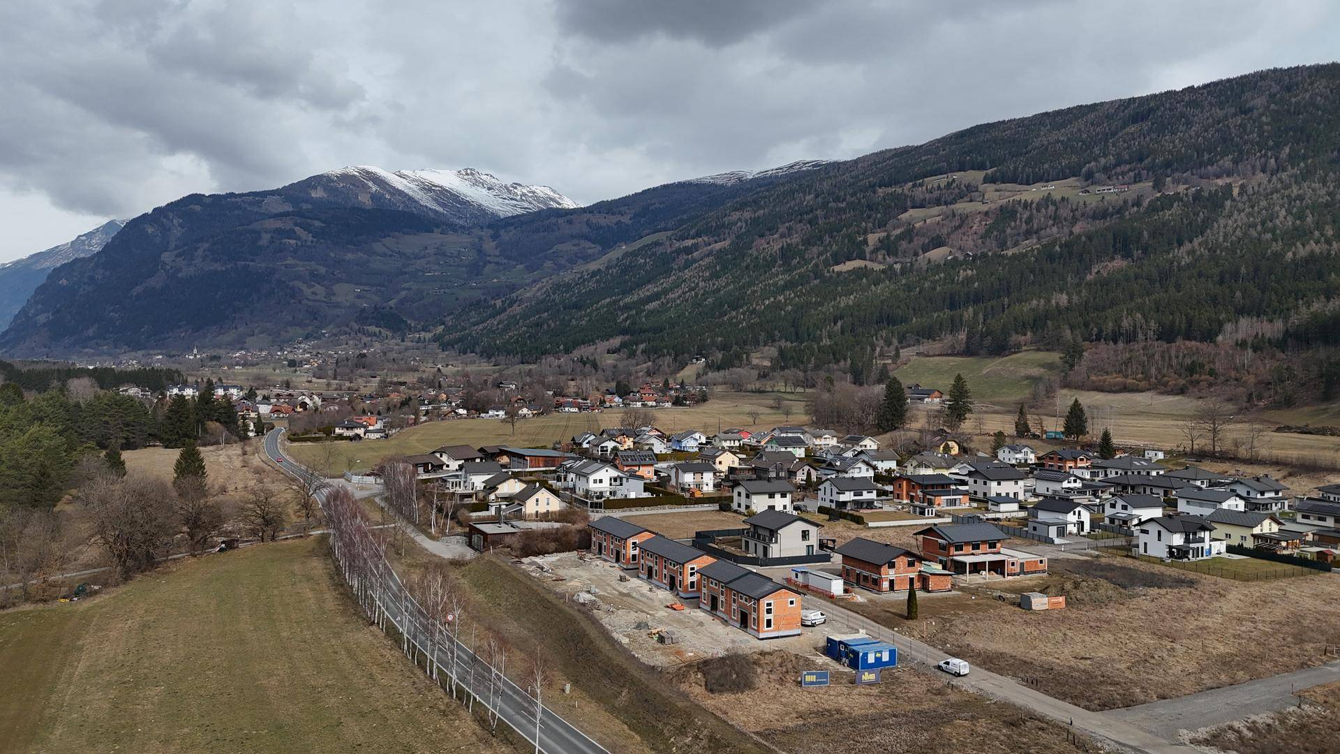 Panoramablick auf eine ländliche Siedlung mit Neubauten und schneebedeckten Bergen im Hintergrund.