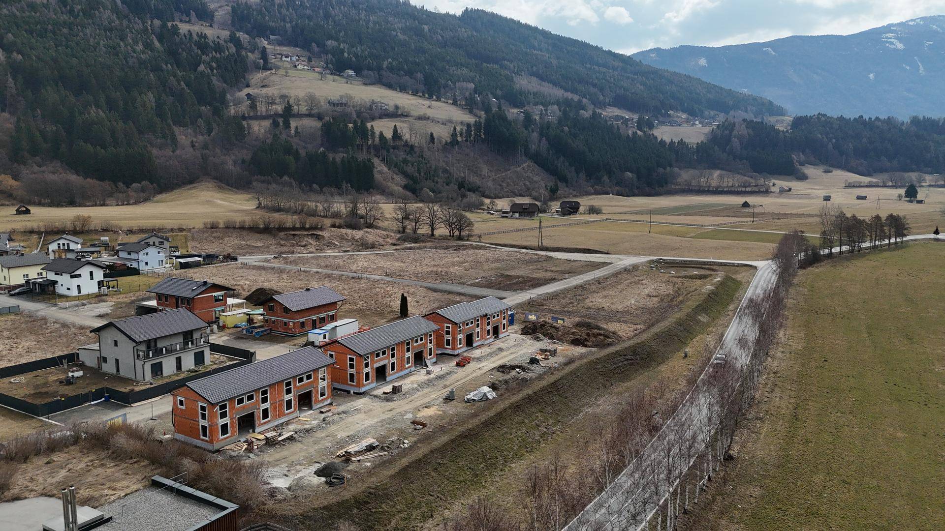 Weitläufige Luftaufnahme der Neubausiedlung in einer grünen Tallandschaft mit umliegenden Bergen.