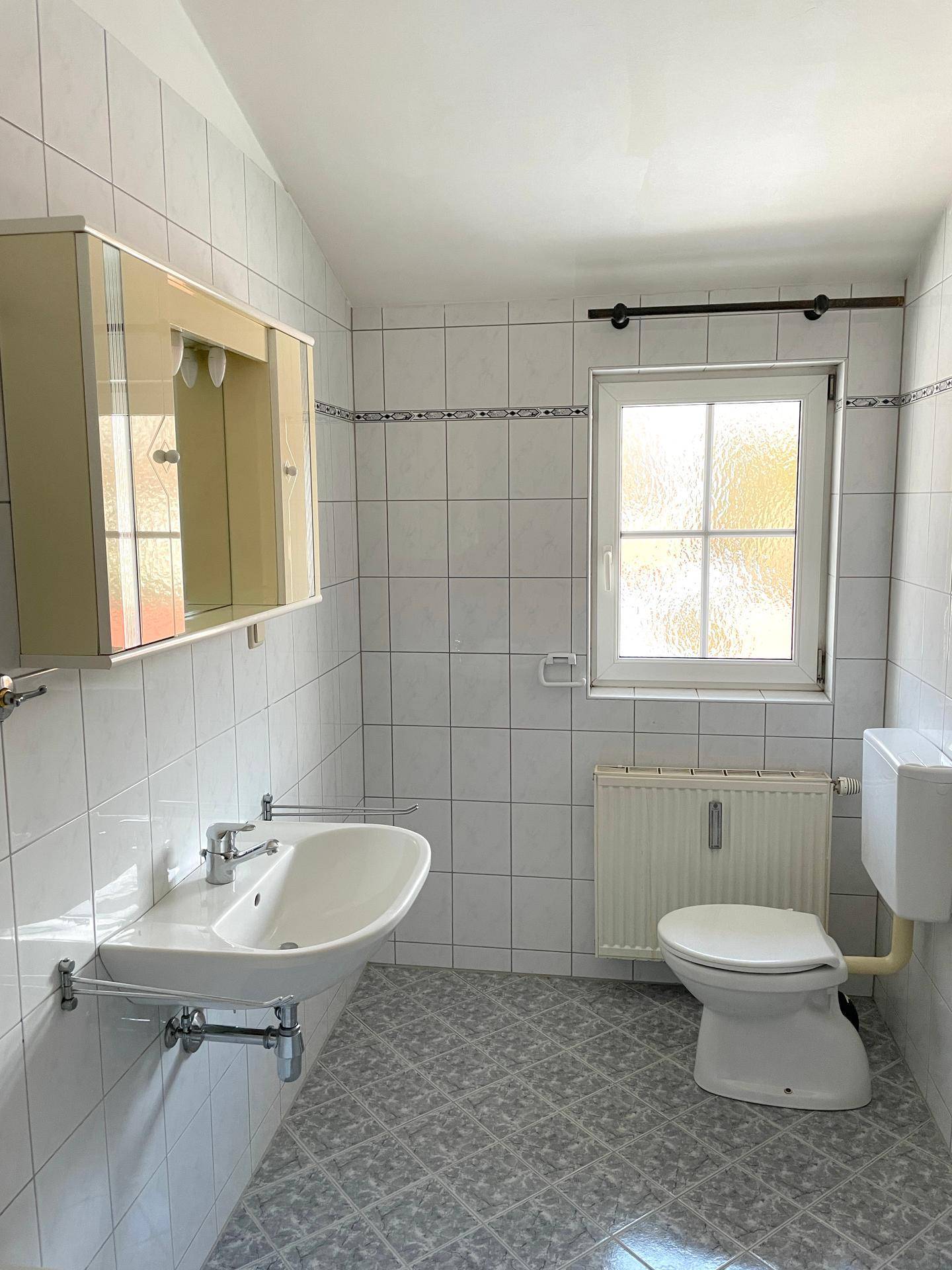 Helles Badezimmer mit weißen Fliesen, Waschbecken, Toilette und einem Fenster für Tageslicht.