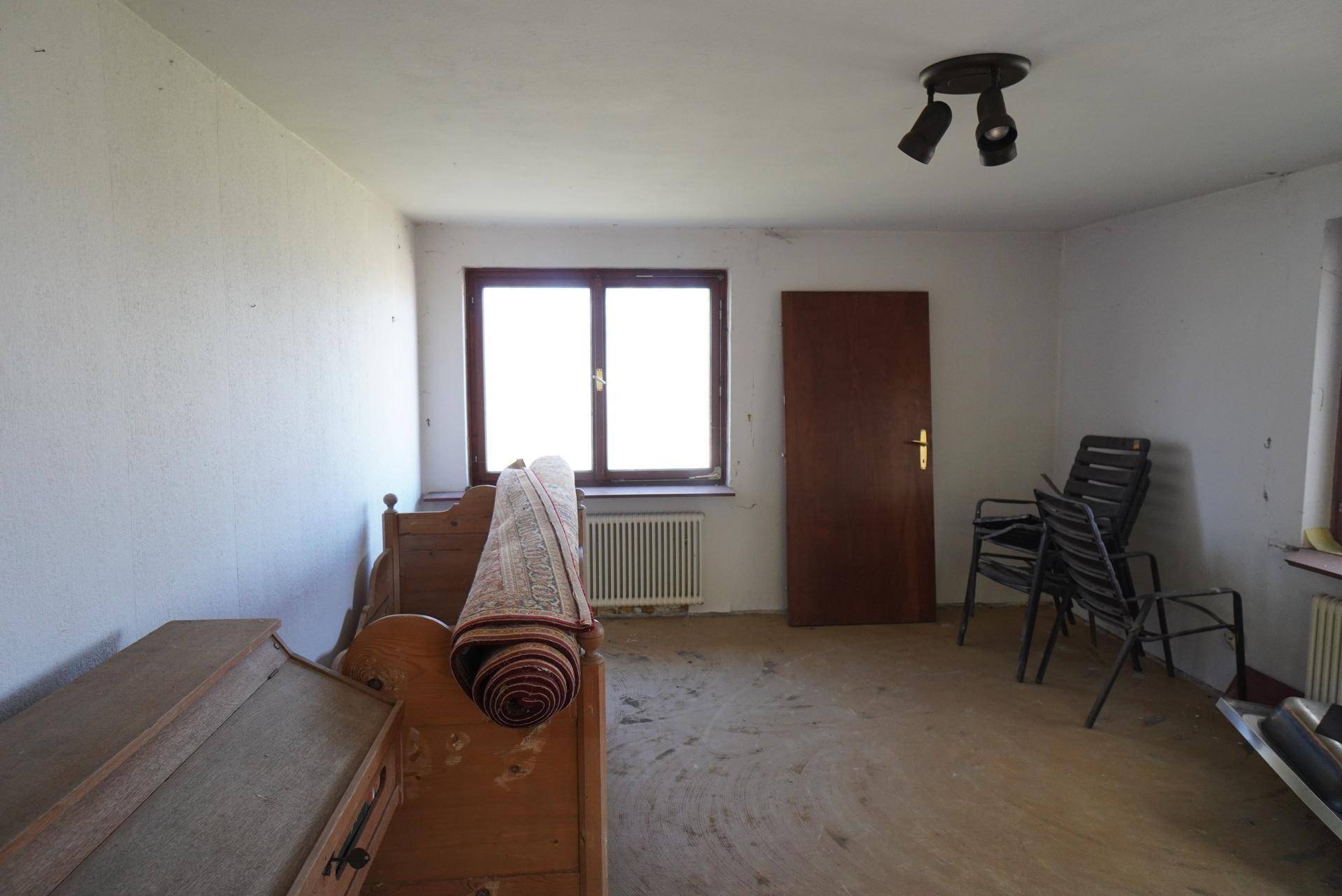 Helles Schlafzimmer mit einem Fenster, einfacher Einrichtung und Renovierungsbedarf.