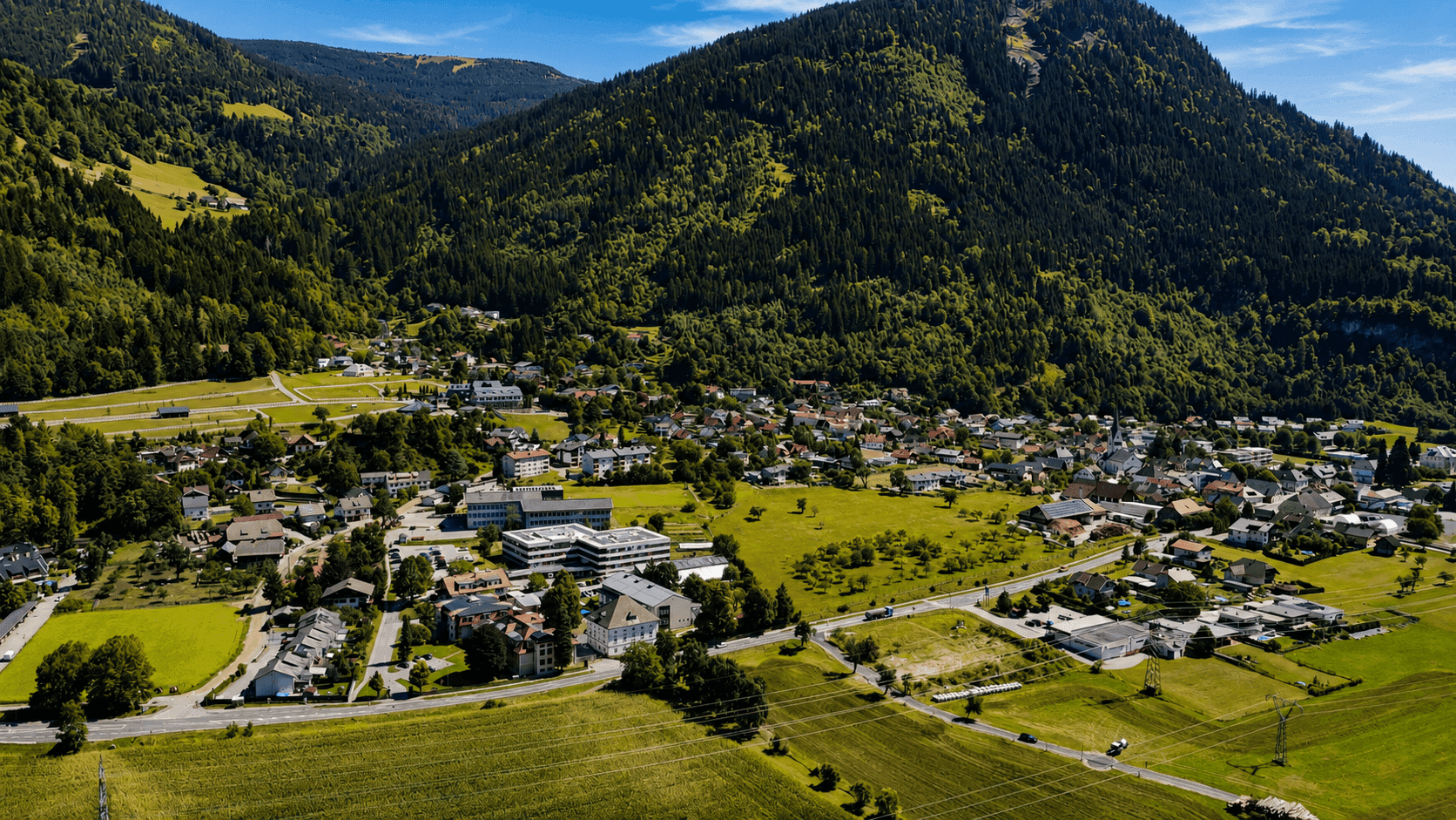 Panoramablick auf ein Tal mit Bergen, einer Ortschaft und landwirtschaftlich genutzten Flächen.