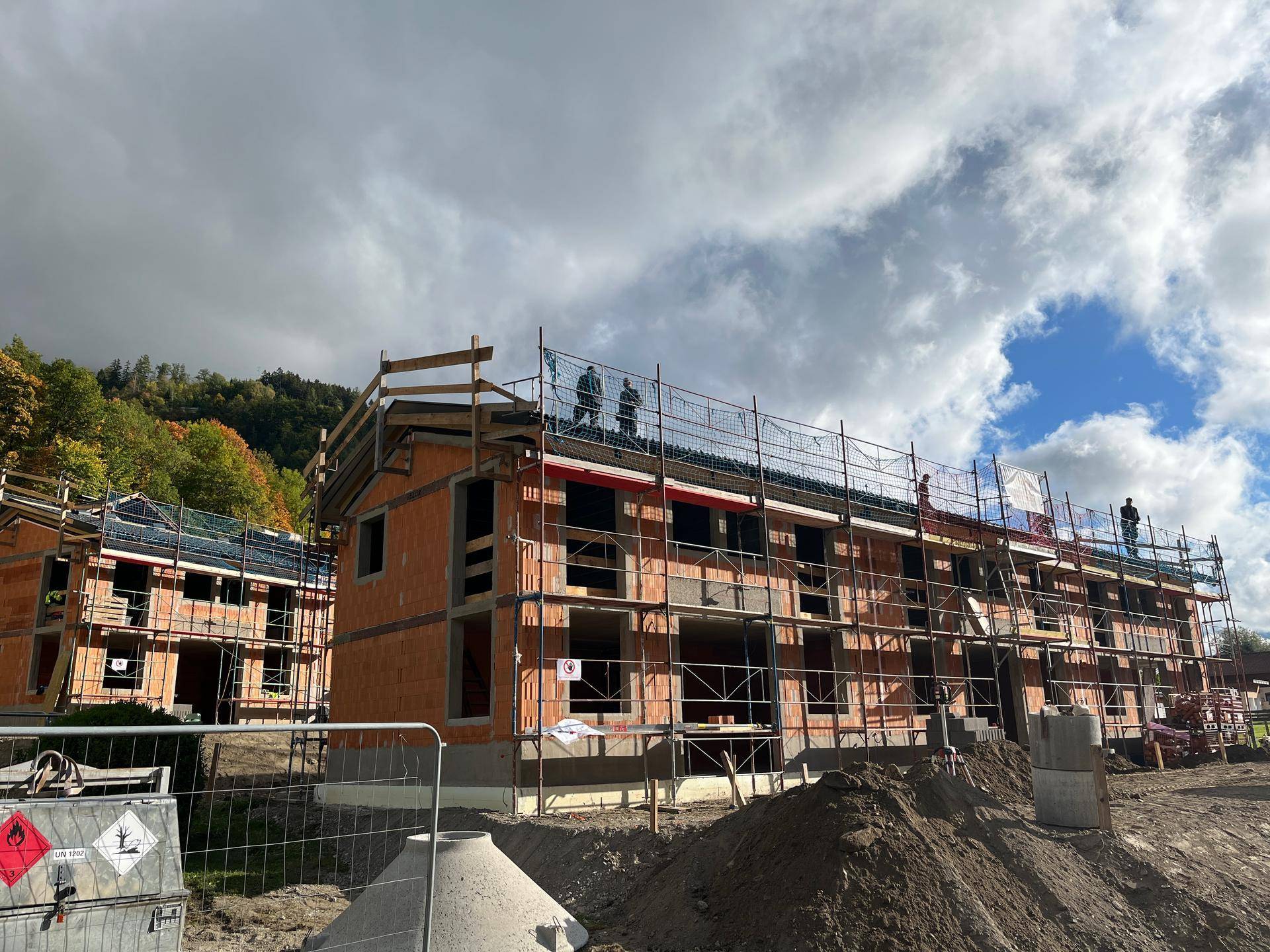 Neubauprojekt mit mehreren Gebäuden im Rohbau, umgeben von Gerüsten und Baumaterialien unter bewölktem Himmel.