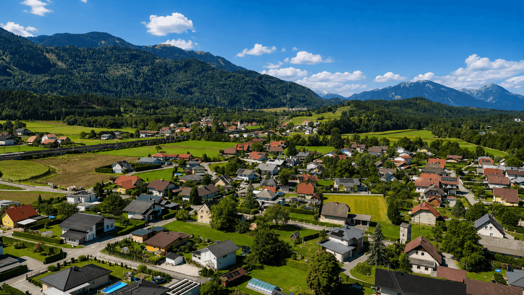 Panoramische Luftaufnahme einer Wohngegend mit vielen Häusern, Feldern und einer beeindruckenden Bergkulisse.